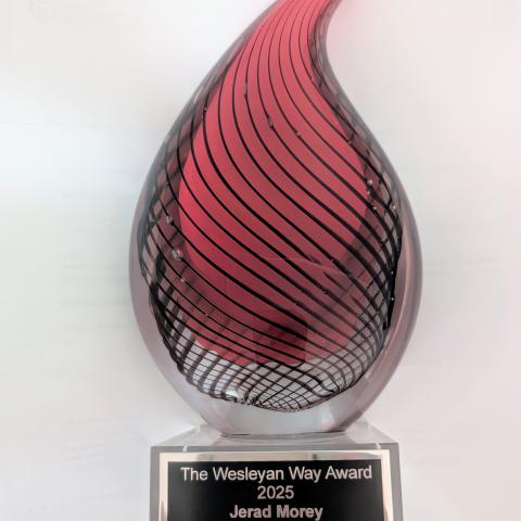 An award in the shape of a flame with a red interior, glass exterior, and a plaque on the bottom reading "The Wesleyan Way Award 2025 Jerad Morey 'Do no harm. Do Good. Stay in Love with God."