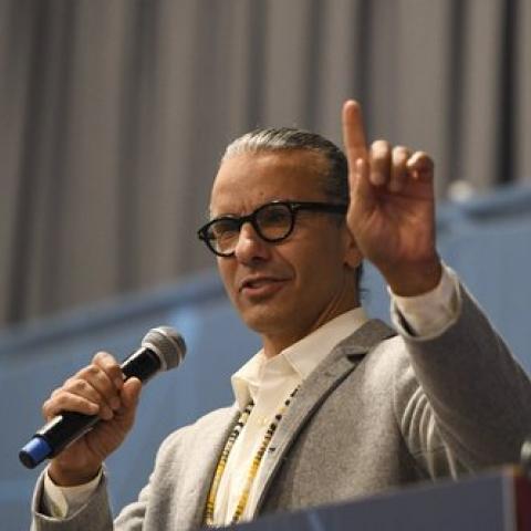 Man with black ponytail and gray blazer stands behind a podium, microphone in one hand and finger raised in another, as though inviting the audience to be patient with a point he's building up to.