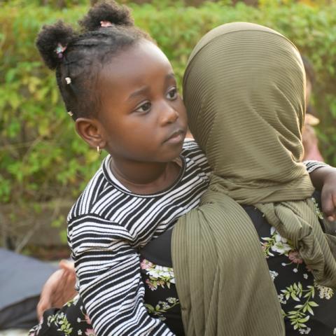 Woman wearing hijab faces away from camera while holding and hugging a young girl, whose open face looks with curiosity past the viewer