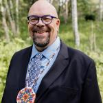 A man in a suit with an indigenour art necklace stands, smiling and looking at the camera, in an outdoor forested setting